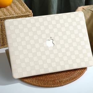 May include: A white laptop case with a checkered pattern and an Apple logo. The case is made of a textured material and has a slightly raised edge around the perimeter.
