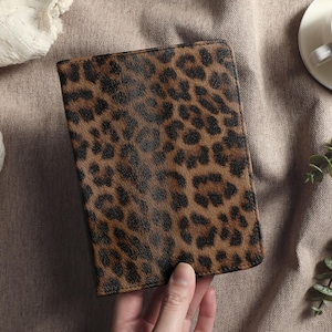 May include: A rectangular, brown and black leopard print wallet. The wallet is held by a hand and is set against a neutral background with a cup of tea and greenery.