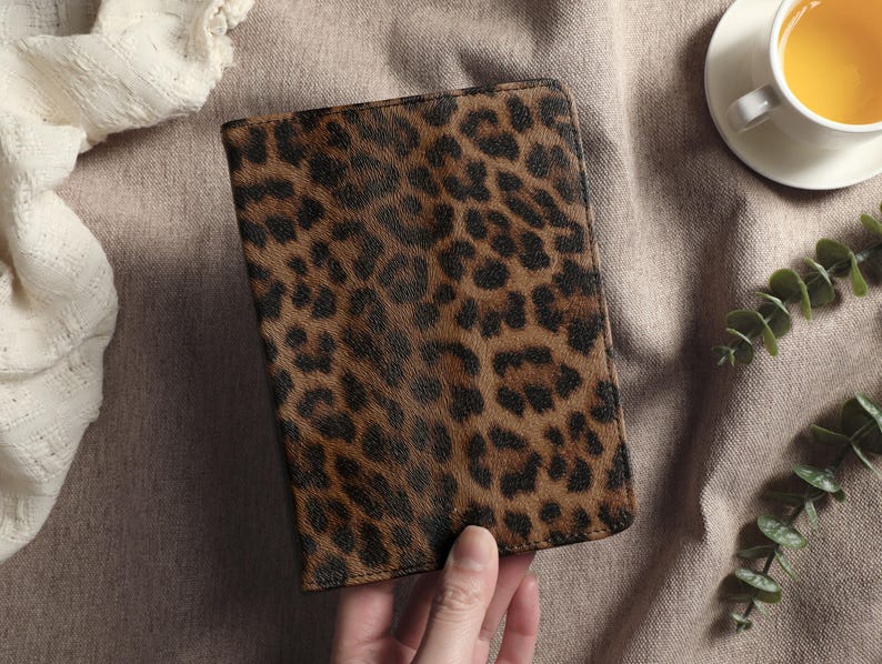 May include: A rectangular, brown and black leopard print wallet. The wallet is held by a hand and is set against a neutral background with a cup of tea and greenery.