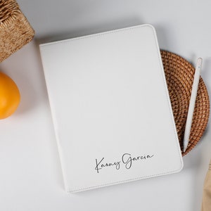 Op de afbeelding: Witte leren tablethoes met de naam "Kanny Garcia" in script. De hoes ligt op een geweven mat met een stylus. Sinaasappels en een rieten mand staan op de achtergrond. Een beige tas met een bruine band staat rechts.