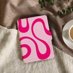 Puede incluir: Una funda rosa para tableta con un diseño abstracto magenta. La funda es rectangular y presenta un patrón repetitivo de líneas curvas. La funda está sobre una superficie texturizada, junto a una taza de té.