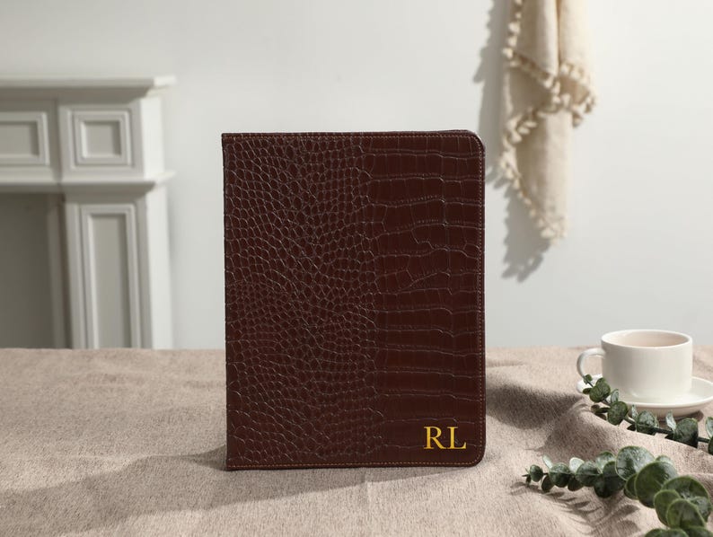May include: A brown leather tablet case with a textured, crocodile-like pattern. The case has a gold-colored monogram with the initials "RL" in the lower right corner. The case is closed and sits on a neutral-colored surface.
