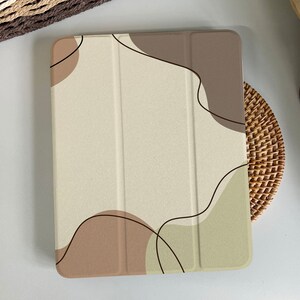 Puede incluir: Una funda beige para tableta con un diseño abstracto con formas marrones y verde oliva delineadas en negro. La funda descansa sobre un salvamanteles de mimbre tejido, con un fondo neutro.