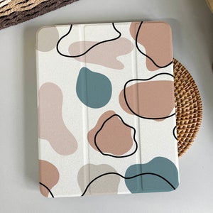 Puede incluir: Una funda blanca para iPad con un patrón de formas abstractas en tonos marrones, azules y beige.