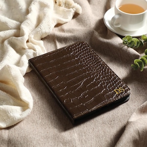 May include: A brown leather journal with a crocodile skin texture, featuring gold initials "DS" in the corner. The journal is on a neutral-toned surface, with a cup of tea and decorative greenery in the background.