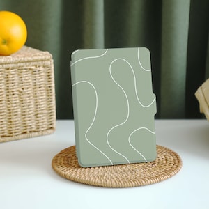 Puede incluir: Un lector electrónico de color verde salvia con un diseño abstracto blanco en la portada. El lector electrónico está sobre un posavasos tejido.