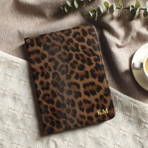 May include: A brown and black leopard print journal with gold initials "KM" on the cover. The journal is closed and resting on a textured white and beige surface. A cup of tea is nearby.