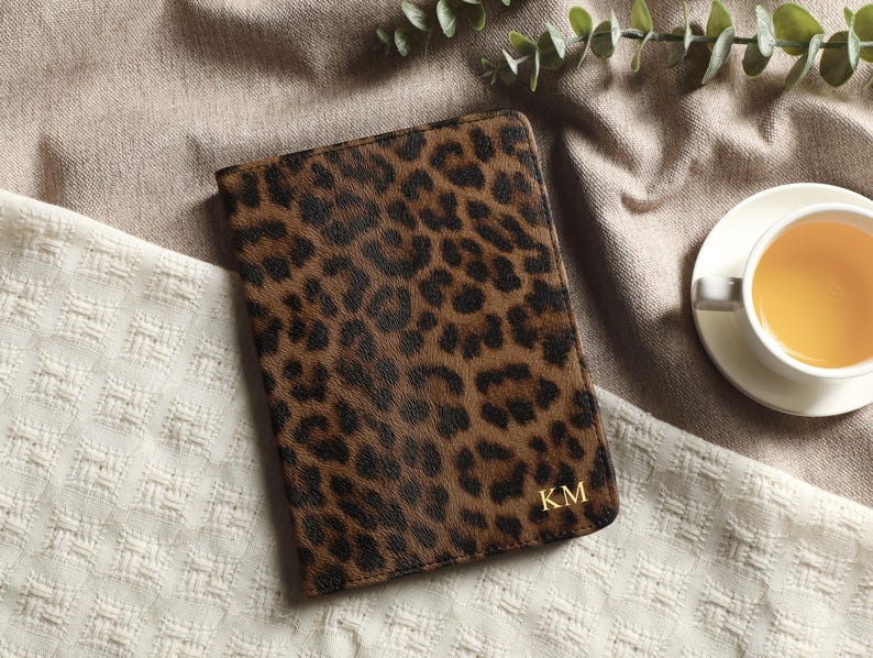 May include: A brown and black leopard print journal with gold initials "KM" on the cover. The journal is closed and resting on a textured white and tan surface. A cup of tea sits nearby.