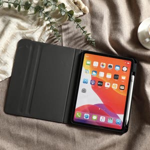 May include: An open black tablet case with a tablet displaying a colorful home screen. A white stylus is attached to the side. A cup of tea sits on a saucer in the upper right corner. The tablet and case are on a brown surface.