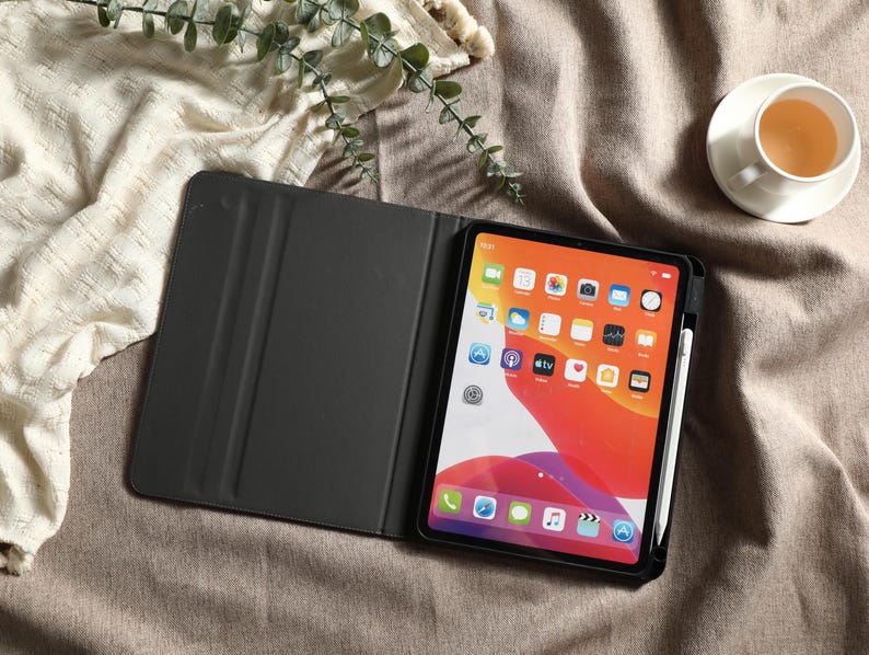 May include: An open black tablet case with a tablet displaying a colorful home screen. A white stylus is attached to the side. A cup of tea sits on a saucer in the upper right corner. The tablet and case are on a brown surface.