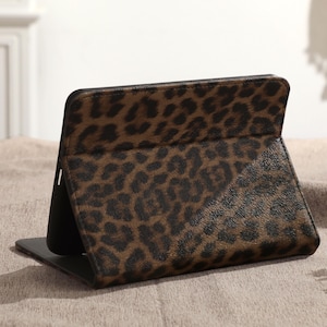 May include: A brown and black leopard print tablet case, standing upright. The case is open, revealing a black interior. The leopard print pattern is a repeating design of dark brown spots on a lighter brown background. The case is on a neutral-colored surface.