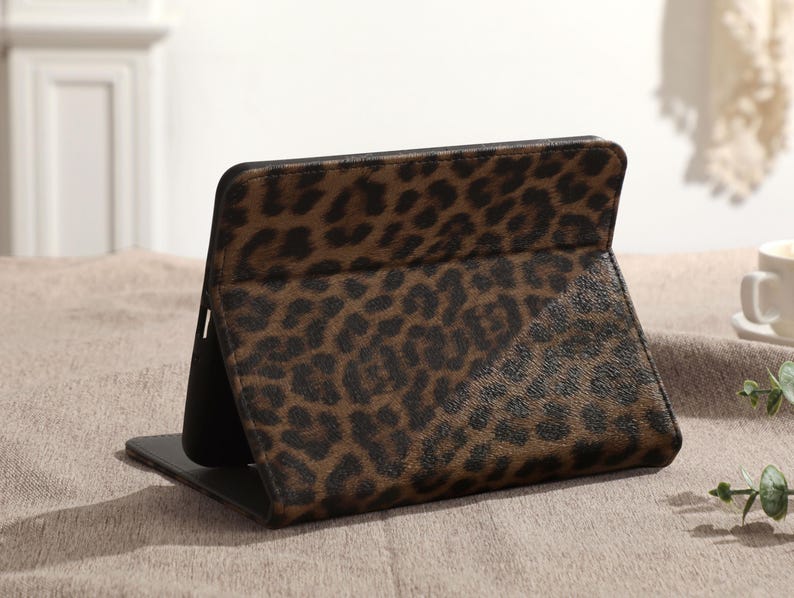 May include: A brown and black leopard print tablet case, standing upright. The case is open, revealing a black interior. The leopard print pattern is a repeating design of dark brown spots on a lighter brown background. The case is on a neutral-colored surface.