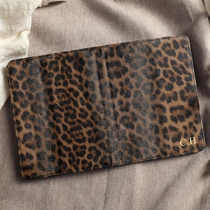 May include: A rectangular, brown and black leopard print passport holder. The passport holder has a gold-colored monogram of the initials "C.H." in the bottom right corner. The passport holder is on a neutral-colored surface.