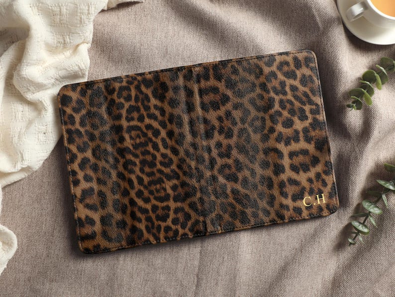 May include: A rectangular, brown and black leopard print passport holder. The passport holder has a gold-colored monogram of the initials "C.H." in the bottom right corner. The passport holder is on a neutral-colored surface.