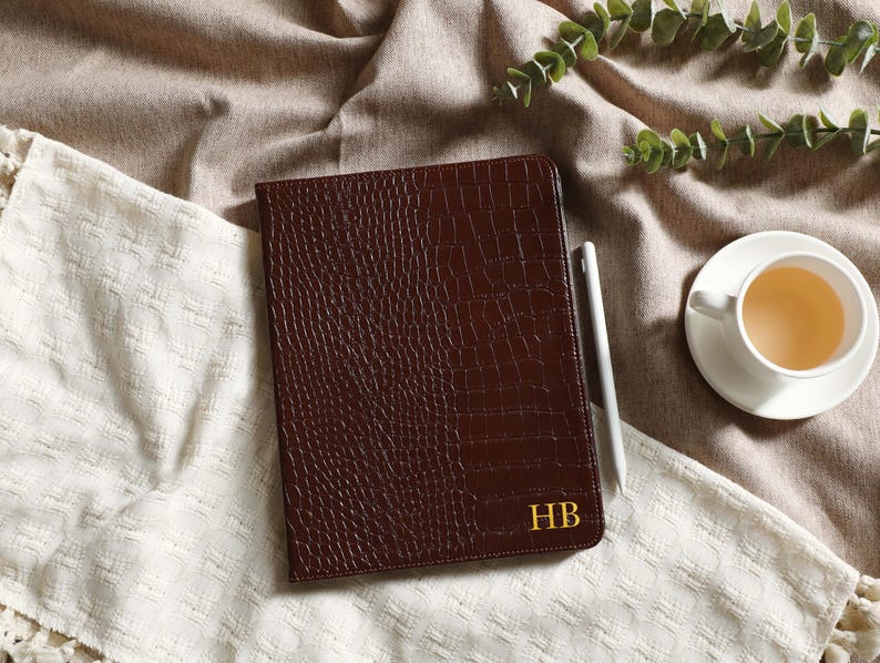 May include: A brown leather tablet case with a crocodile skin texture. The case has gold initials "HB" in the lower right corner. A white stylus and a cup of tea on a saucer are next to the case.