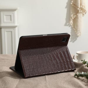 May include: A brown tablet case with a crocodile skin texture is propped open on a table. The case is made of a dark brown material and has a camera cutout. A white coffee cup and saucer are in the background.