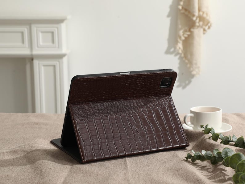 May include: A brown tablet case with a crocodile skin texture is propped open on a table. The case is made of a dark brown material and has a camera cutout. A white coffee cup and saucer are in the background.