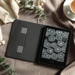 May include: An open black Kindle e-reader with a gray case, displaying a grayscale image of wood cross-sections. The case has a built-in stand. A cup of tea and decorative greenery are also in the image.
