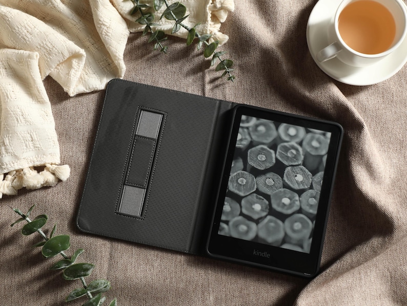 May include: An open black Kindle e-reader with a gray case, displaying a grayscale image of wood cross-sections. The case has a built-in stand. A cup of tea and decorative greenery are also in the image.