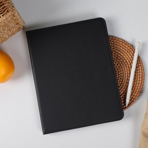 Puede incluir: Una funda de tableta de cuero negro con un lápiz táctil blanco sobre un posavasos tejido. La funda está sobre una superficie blanca.
