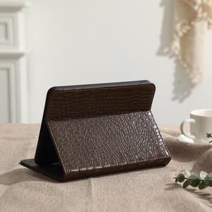 May include: A brown tablet case with a crocodile skin texture is open and propped up on a table. The case is made of a dark brown material and has a black interior. A white teacup and saucer are in the background.