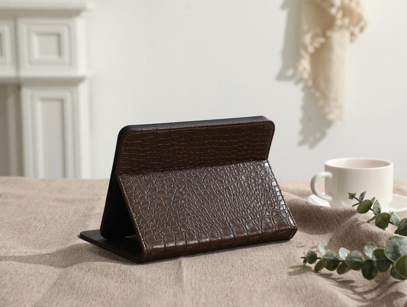 May include: A brown tablet case with a crocodile skin texture is open and propped up on a table. The case is made of a dark brown material and has a black interior. A white teacup and saucer are in the background.