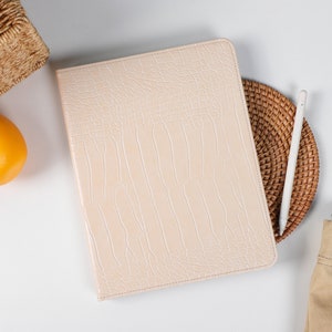 Op de afbeelding: Een licht roze leren iPad-hoes met een gestructureerd krokodillenpatroon. De hoes ligt op een wit oppervlak naast een geweven onderzetter, een witte stylus en een bruine canvas tas.