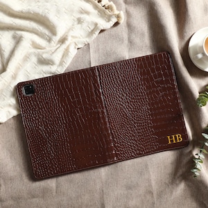 May include: A brown leather tablet case with a textured, crocodile-like pattern. The case is open, revealing the interior. The initials "HB" are in gold on the lower right. A cup of tea and greenery are in the background.
