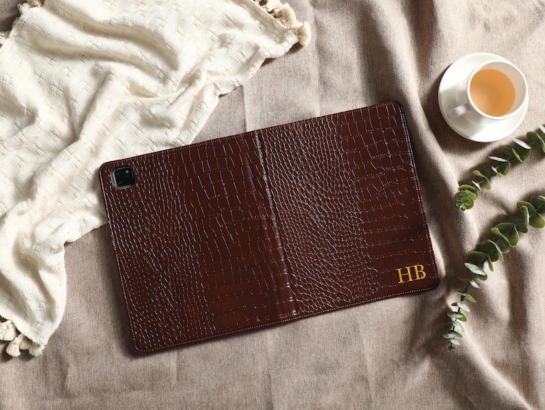 May include: A brown leather tablet case with a textured, crocodile-like pattern. The case is open, revealing the interior. The initials "HB" are in gold on the lower right. A cup of tea and greenery are in the background.