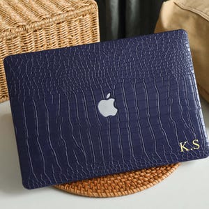 May include: A navy blue laptop case with a crocodile skin texture. The case features a silver Apple logo and gold initials "K.S." on the lower right corner. The case is on a woven mat.