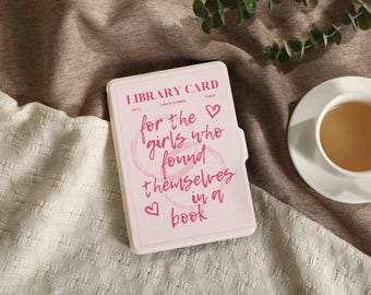 May include: A light pink library card case with the words "LIBRARY CARD" at the top and the phrase "for the girls who found themselves in a book" in pink script. A cup of tea sits on a white saucer.