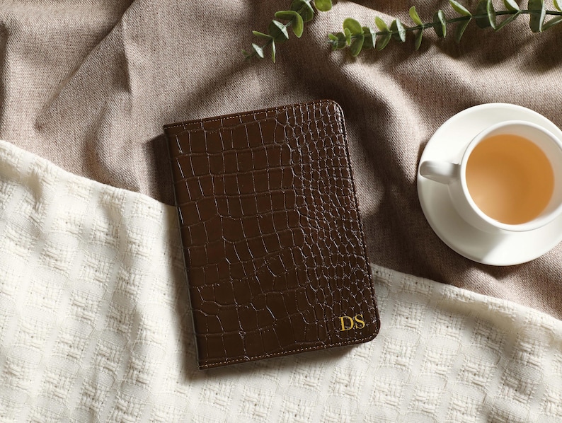 May include: A brown leather journal with a crocodile texture and gold initials "DS" is displayed on a white and brown textured surface. A white teacup filled with tea sits on a saucer nearby, with green leaves in the background.