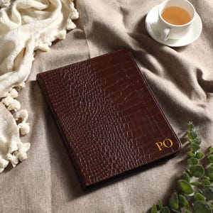May include: A brown leather portfolio with a crocodile skin texture, featuring gold initials "PO". The portfolio is on a neutral-toned surface, accompanied by a cup of tea and decorative greenery. A cream-colored throw with tassels is also present.