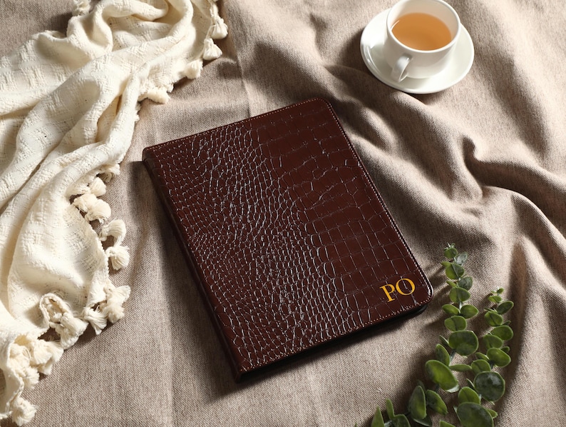May include: A brown leather portfolio with a crocodile skin texture, featuring gold initials "PO". The portfolio is on a neutral-toned surface, accompanied by a cup of tea and decorative greenery. A cream-colored throw with tassels is also present.