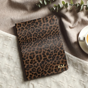 May include: A brown and black leopard print journal with gold initials "KM" is displayed on a textured white and tan surface. A cup of tea sits on a white saucer to the right, with green sprigs in the background. The journal is closed.