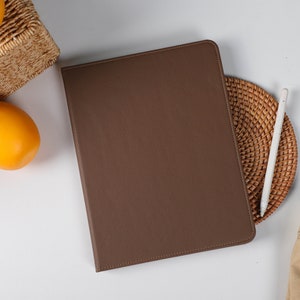 Könnte beinhalten: Eine braune Lederhülle für ein Tablet mit einer strukturierten Oberfläche. Die Hülle ist geschlossen und liegt flach auf einer weißen Oberfläche.