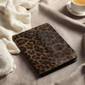 May include: A closed, rectangular journal with a brown and black leopard print cover. The journal is lying on a neutral-colored surface, next to a white, textured cloth and a cup of tea.