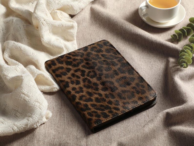May include: A closed, rectangular journal with a brown and black leopard print cover. The journal is lying on a neutral-colored surface, next to a white, textured cloth and a cup of tea.