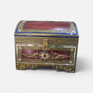 May include: A rectangular wooden chest with a domed lid, featuring a dark red finish, gold-toned filigree metal overlays, blue accents, and small inset mirrors. The front includes a central floral motif, a metal latch, and decorative floral inlays.
