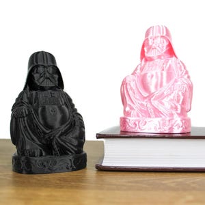Puede incluir: Dos figuras impresas en 3D de Darth Vader como Buda, una en negro y otra en rosa, sentadas en un libro.