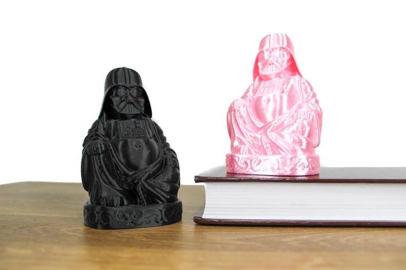 Puede incluir: Dos figuras impresas en 3D de Darth Vader como Buda, una en negro y otra en rosa, sentadas en un libro.