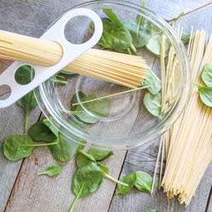 May include: A white pasta portioner with three holes, holding a bundle of cooked spaghetti. A glass bowl with spinach leaves and another bundle of spaghetti are on a wooden surface.