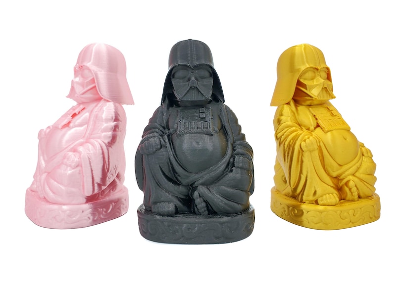 Puede incluir: Tres figuras de cer&aacute;mica de un Buda, cada una pintada en un color diferente: rosa, negro y dorado. Las figuras est&aacute;n todas en la misma pose, con las manos en un gesto de bendici&oacute;n.