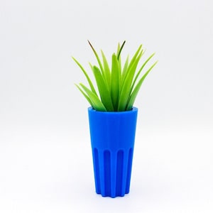 May include: A small, vibrant blue planter with vertical grooves holds a cluster of bright green artificial succulent leaves. The planter has a cylindrical shape and sits against a clean white background. The overall aesthetic is modern and minimalist.