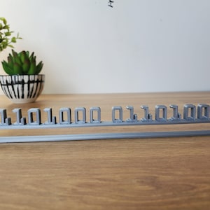Puede incluir: Una pantalla de código binario gris impresa en 3D. Los números "0" y "1" están dispuestos horizontalmente sobre una base curva. La pantalla está sobre una superficie de madera, con una planta en maceta en el fondo.