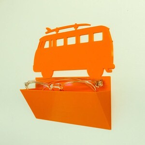 May include: Orange wall-mounted glasses holder shaped like a vintage van with a surfboard on top. The holder has a tray to store glasses. Clear-framed glasses are placed in the tray. The holder is mounted on a white wall.