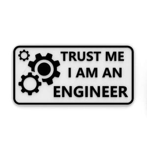 May include: A rectangular white sticker with a black border. The sticker features three black gear icons and the text "TRUST ME I AM AN ENGINEER" in a bold, sans-serif font. The design is simple and direct.