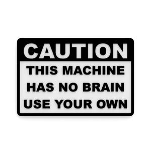 May include: A rectangular black and white sign with the word "CAUTION" at the top. Below, the text reads "THIS MACHINE HAS NO BRAIN USE YOUR OWN". The sign has a white background and a black border.