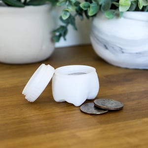 May include: White ceramic tooth-shaped container with a lid, sitting on a wooden surface with three silver coins beside it.