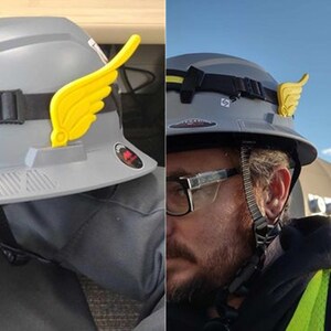 May include: A gray hard hat with a yellow wing-shaped accessory attached to the side. The hard hat has a black strap and a white sticker with the word "STORM" on it.